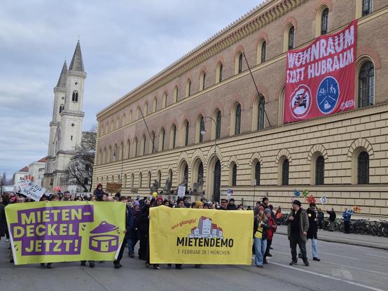Foto der Mietendemo vom 07.02.26 in München, Ludwigstraße (Höhe Bayerische Staatsbibliothek). Demonstrierende halten Banner "Mieten-Deckel Jetzt" und "uns glangts" - dahinter ein Bannerdrop von XR München "Wohnraum statt Hubraum" an der Fassade der Bayerischen Staatsbibliothek.