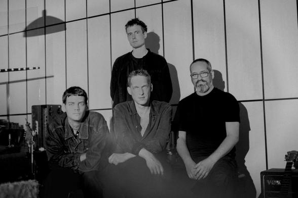 Ein Band-Photo in scharz-weiß.
Vor einer weißen Wand, die durch schwarze Linien in vertikale Rechtecke unterteilt ist, sitzen drei Musiker (ohne Instrumente). Hinter dem Musiker in der Mitte steht ein vierter Musiker.
Am rechten unteren Rand sind ein Verstärker samt Gitarre zu erkennen (angeschnitten).
Links oben befindet sich der Schatten einer hängenden Lampe.
Darunter ein Schlagzeug (angeschnitten) und ein weiterer Verstärker.