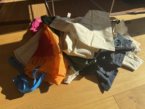 A photo of a pile of about 20 fabric shopping bags in different colors. Most of them have the standard size, they are on a wooden floor with a coffee table in the background. The sun is shining in from the right.