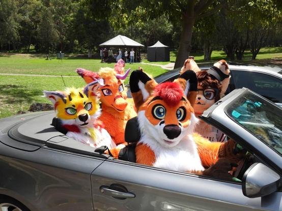 Colto Fox in the driver's seat of a grey/silver BMW convertible. He's accompanied by Kyle next to him, Briggs and Lino Plum in the rear seats. They're parked outside of a park with some gazebos in the distant background with many people around them.
