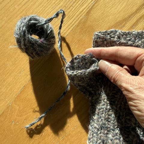 Das nun fertig verzwirnte und nochmal gezwirnte Garn liegt in einem kleinel Knöuel wiederum auf dem hellen Holzboden in der Sonne. Daneben liegt jetzt der fingerlose Handschuh, dessen Daumenabschluss abgenutzt ist und repariert werden muss. Ich halte die Stelle in meinen Fingern.