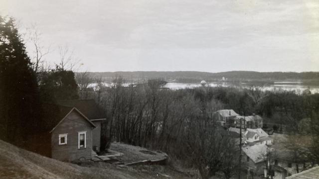 A black and white photo shows a view of the Mississippi River from the bluff at Fort Madison, Iowa.  In the foreground, a steep slope with one small house nestled into the hillside.  Below, the houses in the town.  The river itself is a silvery strip. Photo from early 1980s.