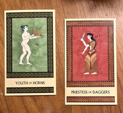 Two Minoan Tarot cards side by side on a brown wood surface. The Youth of Horns, on the left, is painted in shades of green and brown and has an intricate rectangular border. It shows a naked Minoan boy standing, facing right, holding a pair of tan sacred horns in front of him. The Priestess of Daggers, on the right, is painted in shades of deep red and tan and has an intricate rectangular border. It shows a Minoan woman in a long dress, standing, facing left, brandishing a dagger in front of her.