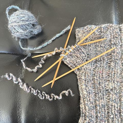 Rechts im Bild der fingerlose Handschuh. Er sieht unregelmäßig aus, weil das verwendete Garn aus wild gemixten Resten gesponnen wurde. Die Farbe ist hellgrau-grünlich mit weißen und pinkfarbenen Stellen, kurz: die hässlichsten fingerlosen Handschuhe der Welt. Aber auch die besten, und das seit 2008.
Der Abkettrand am Daumen wurde aufgepfriemelt (Fachausdruck) und zwei weitere Reihen rückwärts gestrickt, was eine gute Idee war, weil ich so noch zwei weitere dünne Stellen beseitigen konnte.
Die Maschen liegen auf einem Bambus-Nadelspiel, das neue Garn für die Reparatur liegt als kleines Knäuel links oben im Bild.