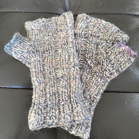 Beide Handschuhe liegen auf schwarzem Leder-Hintergrund. beim rechten Handschuh ist der Abkettrand am Daumen bereits von zwei Jahren repariert worden mit pink-lila Garn. Der Daumenabschluss des linken Handschuhs ist gerade repariert worden und ist nun grün-türkis.