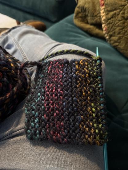A dark blue yarn intermingled with rainbow colors, knit into the beginning of a scarf, lying across a jean-clad leg.