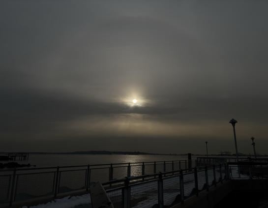 A view from a pier overlooking the waters of a wide bay. A halo of pale light arcs high over the setting sun as it shines over a strip of dark clouds. Below, golden crepuscular rays fan out in a luminous band before being obscured by more gray clouds at the horizon. Directly beneath the sun, a column of sunlight sparkles on the water. In the foreground are guardrails flanking the pier’s snow-covered walkway as it recedes to some lampposts at right frame.