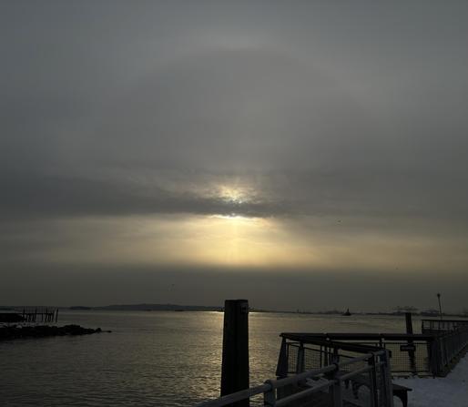 Another view of the sunbow over the pier. The strip of gray clouds obscures the sun itself, without diminishing the halo above or the crepuscular rays below — a strikingly dramatic effect. Directly below the sun is the silhouette of a wooden pillar rising out of the bay, surrounded by the glow of the sun sparkles on the water.