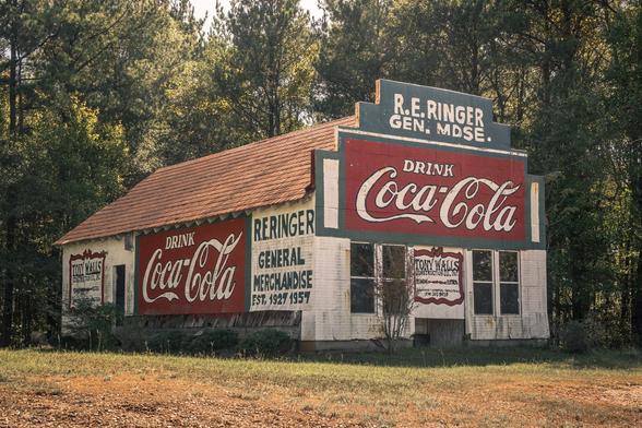 Abandoned white wooden general store with rusted corrugated metal roof, featuring multiple vintage Coca-Cola advertisements painted on exterior walls. Sign reads "R.E. RINGER GEN. MDSE." and "R.E. RINGER GENERAL MERCHANDISE EST. 1927 1957." Building stands in overgrown field with dense forest background.