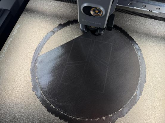 Overhead view down into a BambuLab H2S 3D printer as it lays down a smooth large disc of black flexible filament on its textured printing plate.