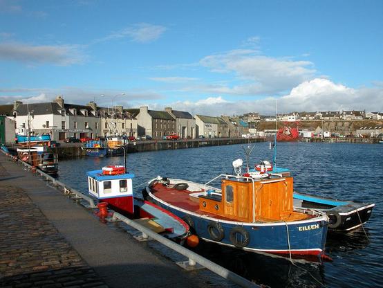 The harbour at Wick in Caithness. The image shows a broad harbour with a quayside in the foreground on the left, with small boats moored at it. In the background the harbour is lined by mainly three storey buildings. The scene is in sunshine.