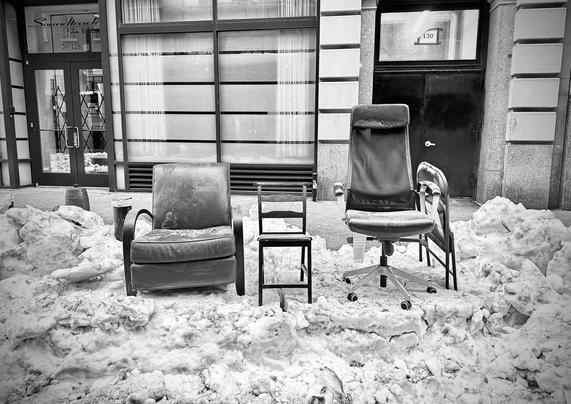 A black and white photo taken from the street at a 3 feet tall line of snowy ice by the sidewalk. On top of the snow are lined up four chairs. The one on the left is a big leather easy chair with rounded arms that looks modern. The next one is a small wooden straight back chair that looks like it's for a child. The next chair is an office chair on wheels with an extremely high back. And leaning on that chair is a regular metal folded chair. Behind them are windows and doors from a nondescript building