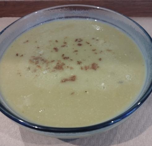 Una crema o sopa de color beige verdoso claro se presenta en un tazón de vidrio transparente. La superficie de la crema está espolvoreada con pequeñas partículas marrones, que parecen ser especias molidas o semillas. El tazón descansa sobre una superficie textil clara.