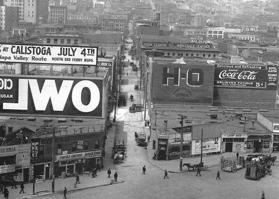Black and white photo taken from an elevated position. It shows an urban setting with large buildings either side of a road that runs from the foreground to the background. Some of these buildings have advertisements painted on them, and the most prominent of these has white lettering out of a dark background that reads "Owl" (from Owl Cigars) but written in reverse.