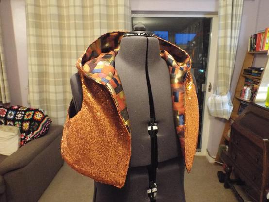 A cropped sleeveless hoodie on a mannequin. The garment is completed in an orange sequined fabric on the outside, and a metallic brocade on the inside.