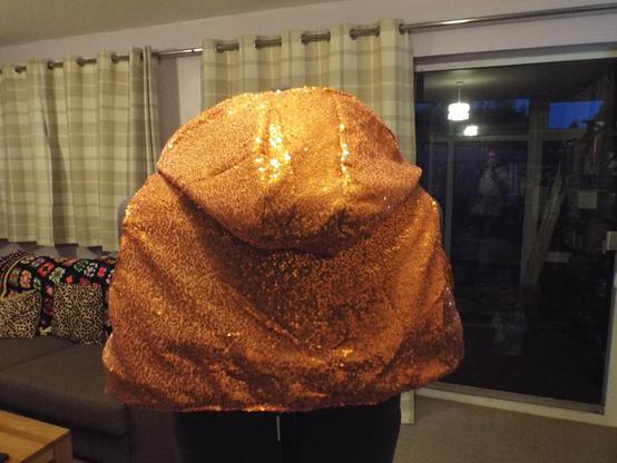 A cropped sleeveless hoodie on a mannequin. The garment is completed in an orange sequined fabric on the outside, and a metallic brocade on the inside.