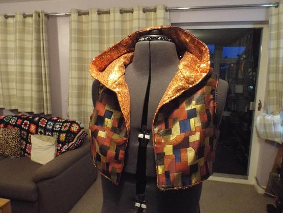 A cropped sleeveless hoodie on a mannequin. The garment is completed in an orange sequined fabric on the inside, and a metallic brocade on the outside.