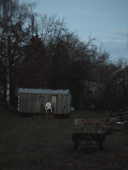 A weathered caravan with a white chair outside, surrounded by trees and scattered items in a dark, outdoor setting at dusk.