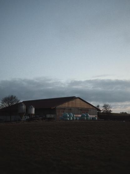 A barn with hay bales wrapped in blue plastic in front, set against a cloudy sky during dusk.