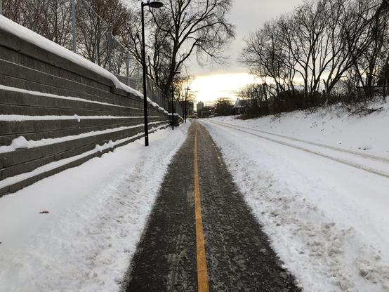 A multi-use path recedes into the distance. There is snow on the ground on either side of the path. On the left there’s a retaining wall. On the right there’s a railroad track. There are bare trees on both sides. The sky is grey with clouds.