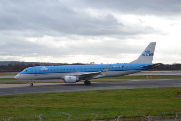 Side view of a twin engined jet airliner taxiing from right to left along a grey taxiway.
The plane is light blue on the top, with a white belly, and a thin, darker blue stripe running along the body separating the 2 coloured sections.
There are white "KLM" titles under a stylised white crown on the upper forward fuselage, a design reversed and repeated, blue "KLM"  under a blue crown on the sides of the engine pods under the wings, and in a larger form on the white tail.
A large expanse of grass fills the foreground, lining the side of the taxiway, with more grass divided by more taxiways and a runway in the background.
Dark green trees meet rather dramatic cloudy sky in the distance.