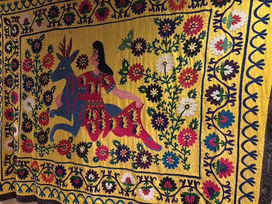 A vibrant Uzbek Suzani textile art piece featuring a woman in traditional clothing sitting beside a stylized blue deer, surrounded by colorful floral motifs on a bright yellow background.