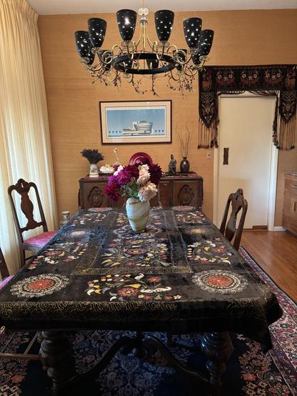 A dining room featuring an ornate table covered with a decorative black Uzbek Suzani adorned with floral patterns. A vase of colorful flowers sits in the center. The room has elegant wooden furniture, a chandelier with black shades, and a piece of artwork on the wall.