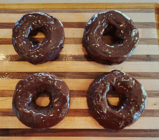 Four chocolate donuts, covered in a chocolate glaze, resting on a wooden cutting board. They want to be eaten. Mmmmm donuts...