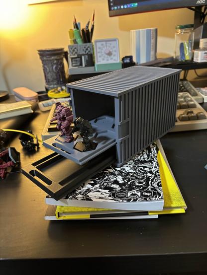 A close-up view of a desk featuring a model shipping container propped open, with Battletech Mechs placed inside I t. Various stationery items, a timer, and a pen holder are visible in the background, along with a couple of TTRPG books stacked underneath.