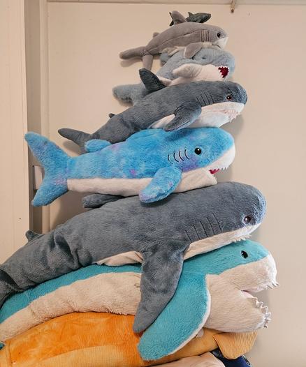 A pile of plush sharks, arranged from bottom to top in decreasing size.  The bottom shark is slightly bigger than a blahaj plushie, and the top shark is a tiny plastic whale shark. There are 7 sharks all up.  The pile is set upon a partially visible giant squid plushie