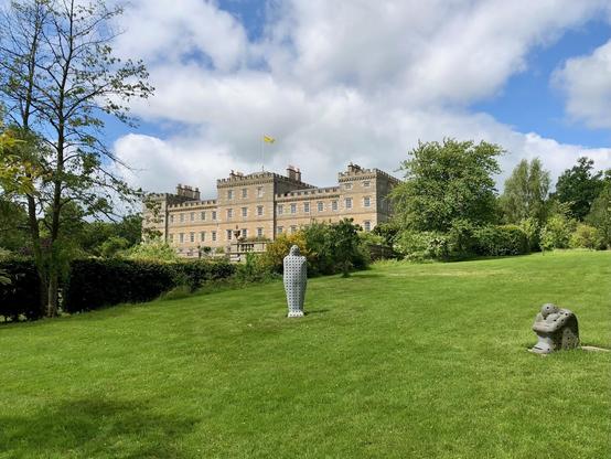 A large rectangular stone building with crenellated parapets stands in the background, topped by multiple chimneys and a yellow flag. The structure, with evenly spaced sash windows, is surrounded by lush greenery and landscaped gardens. In the foreground lies a neatly maintained lawn with two modern outdoor sculptures: a tall white perforated figure and a smaller grey abstract piece. Trees and hedges frame the scene under a partly cloudy sky with patches of blue, giving a sense of open space and parkland.