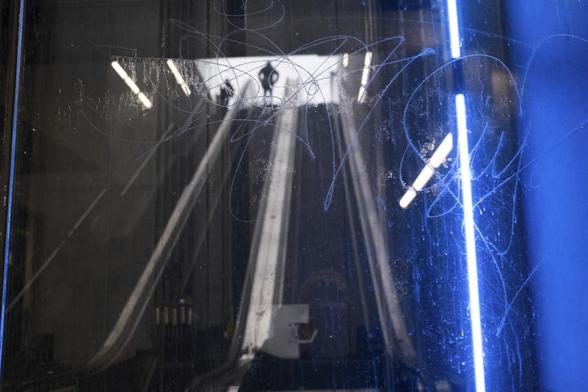 A man on the top of an escalator, the hands on his hips is about to go down into a metro station In Brussels. His black silhouette contrasts with the framed of light at the top of the staircase. The image, seen through the scratched window of an elevator is dominated by grey and dark tones that contrasts with an electric blue part illuminated by a neon.