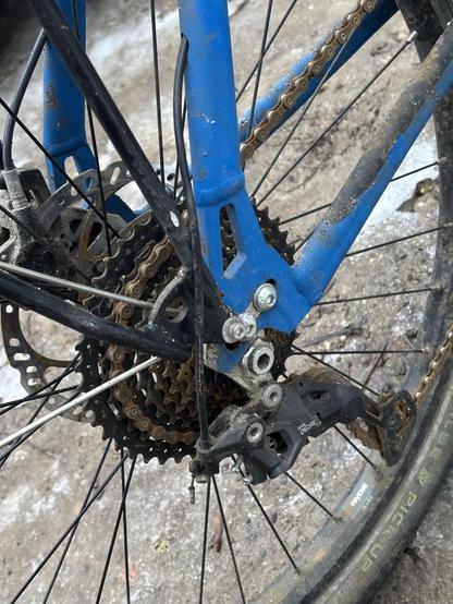 Nahaufnahme des Hinterrads eines Fahrrads, das den Umwerfer, die Kassette, die Kette und einen Teil des Rahmens zeigt. Die Komponenten sind mit Schmutz bedeckt, was auf die jüngste Nutzung unter Bedingungen im Freien hinweist.