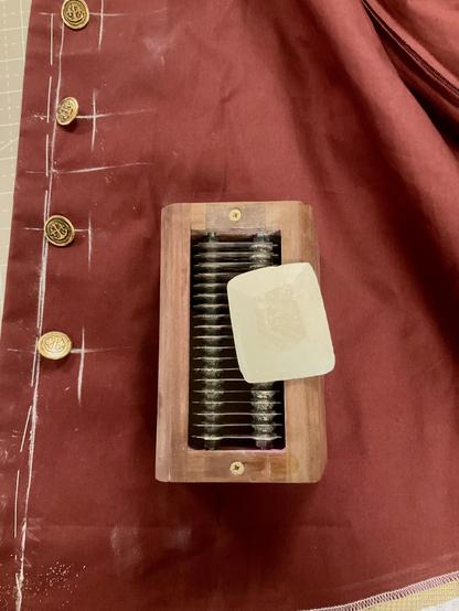 Auf einem Rostroten Baumwollkanvas sind goldene Knöpfe ausgelegt und Knopflöcherpositionen mit Kreide markiert. Im Vordergrund steht eine Box aus Walnussholz mit Rasierklingen an der Oberseite, die Box dient dem schärfen von Schneiderkreide. Die Schneiderkreide liegt auf der Holzbox