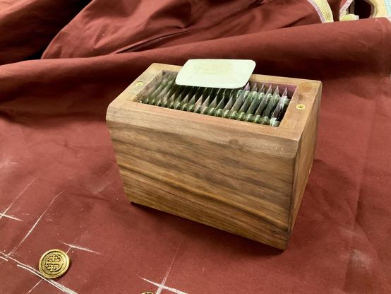 Auf einem Rostroten Baumwollkanvas sind goldene Knöpfe ausgelegt und Knopflöcherpositionen mit Kreide markiert. Im Vordergrund diagonal steht eine Box aus Walnussholz mit Rasierklingen an der Oberseite, die Box dient dem schärfen von Schneiderkreide. Die Schneiderkreide liegt auf der Holzbox