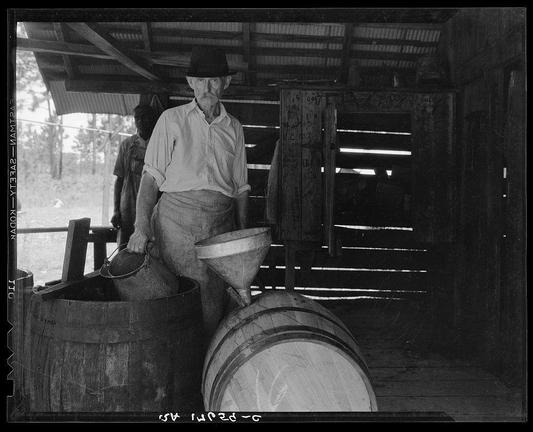 The black and white photograph depicts a man wearing a wide-brimmed hat, sleeveless shirt with rolled-up sleeves, and what appears to be an apron or utility trousers. He is standing at the entrance of a rustic structure made from wood beams, likely part of a larger outdoor setup given glimpses of trees in the background through gaps between wooden planks. In front of him are various tools indicative of manual labor: a large cylindrical barrel that he seems to be interacting with using his right hand; a funnel-like device placed atop another barrel; and what might be a metal bucket or container being held by his left hand, suggesting some sort of processing activity involving liquids. The overall scene suggests an agricultural setting where the man is engaged in laborious work, possibly related to food preparation given historical context implied by attire and tools used.