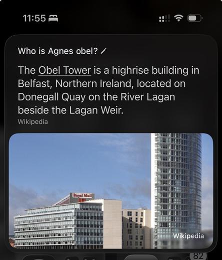 The image shows a tall modern high-rise building, the Obel Tower, in Belfast, Northern Ireland, with its sleek glass façade dominating the right side of the frame. In front of it, there is a shorter rectangular building with a “Royal Mail” sign on top, along with other mid-rise structures clustered around the cityscape. The sky is clear with light clouds, providing a bright backdrop to the urban skyline. This alt-text was autogenerated using Apple Intelligence Private Cloud Compute.