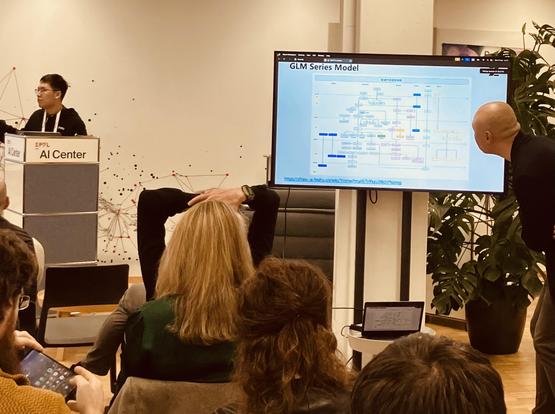 A person presents while another inspects a highly detailed slide, another person in the audience is holding hands over head