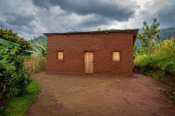 A single-story mud house with a wooden door and two small windows, surrounded by greenery and a dirt path. The sky is overcast with clouds, and hills are visible in the background.