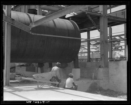 The black and white photograph captures a moment from an industrial or construction setting, showcasing two individuals amidst large machinery. The central figure is seated on the ground in front of what appears to be part of a turpentine still, identified by visible lettering that hints at its function. This individual wears light-colored clothing with sleeves rolled up, typical attire for manual labor during hot weather conditions suggested by their hats and outdoor setting.

To the left stands another figure beside an empty wheelbarrow, dressed in lighter-toned work clothes indicative of a worker engaged in physical tasks within this environment. The machinery they are attending to is large, cylindrical, with riveted seams, supported on metal structures suggesting it's part of larger industrial equipment.

The surrounding architecture includes visible steel beams and concrete supports indicating an unfinished or under-construction setting. A hint of greenery suggests the presence of trees in proximity to the site, contrasting the mechanical environment with natural elements. The overall impression is one of a labor-intensive workspace during what could be early 20th-century industrial practices.

The photograph's monochromatic color scheme and graininess convey a sense of age and historical context, likely from an era when such manual operations were commonplace in ind [...]