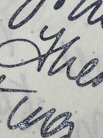 A close-up of glittery, cursive handwriting on a textured surface, featuring dark ink with iridescent shimmer and small blue dots in the background.