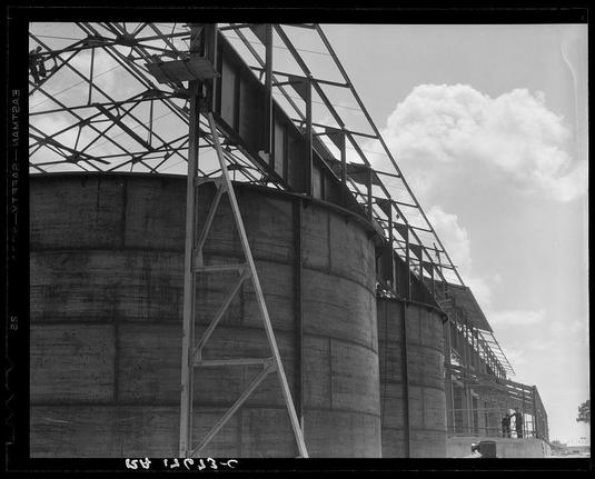 The image is a black and white photograph depicting an industrial scene, showcasing large cylindrical tanks with visible metal framework. These structures appear to be part of a larger complex or factory setting under construction or maintenance. The sky above has scattered clouds indicating fair weather conditions during the time when this photo was taken.
On closer inspection, one can observe what appears to be lumber and scaffolding around parts of the tank structure, hinting at ongoing work such as reinforcement or installation processes. There are two individuals in view; they seem small compared to the large structures surrounding them which emphasizes their scale. The person on the right is standing near a railing, possibly observing the site's operations.
In terms of textual information within the image, there appears to be handwritten text at the bottom left and top areas that might indicate location or context details related to this industrial scene but these are not clearly discernible in the provided description.