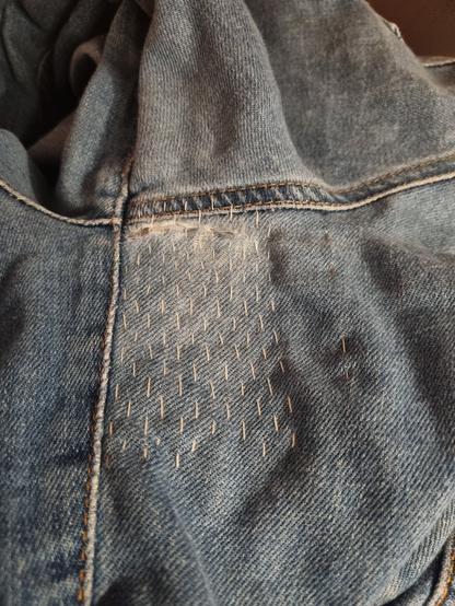 Eine blaue Jeans mit einem Riss in Nähe einer Naht. Auf dem Stoff sind streifenförmige weiße Reparaturstiche zu erkennen