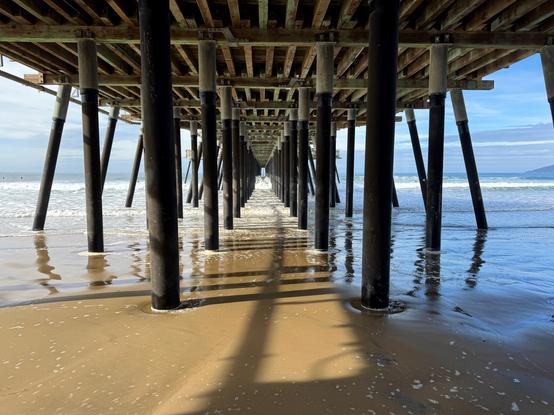 Looking through the repetitive framing of the pier's underside, both the pilings and boards that support the deck of the pier. The pilings look smaller and smaller towards the end of the pier. Waves are rolling in, but they are some distance away since the tide is out. The sun is illuminating the left side of the pier so there is a yellow hue. On the right side, there is a blue hue due to the shadows from the pier. The sky is mostly blue with some thin clouds.