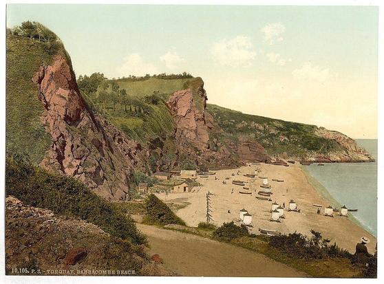 This image depicts a picturesque coastal scene with red and brown cliff faces rising steeply from the shore. The shoreline is sandy, populated by small beach huts or tents arranged in groups along the sand. Several boats are visible on the water near the cliffs and docked at the shore. A dirt path leads down to the beach area through rocky outcrops and greenery that appears lush against the earthy tones of the cliffside rocks.

In the foreground, a figure dressed in period attire sits by themselves on a higher vantage point overlooking the beach, suggesting leisure or contemplation of the view. The overall atmosphere suggests an idyllic seaside setting from approximately late 19th to early 20th century England. Notable for its serene ambiance and natural beauty, this location is indicative of Victorian era seaside resorts popular among British citizens seeking relaxation away from urban life.

The image carries historical significance as a representation of leisure during that period in Torquay's Babbacombe Beach area, known today as one of the most beautiful places to visit on the Devon coast.