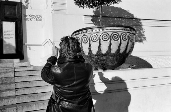The black and white image shows a person in a leather coat standing on steps outside a building, raising their hands as if framing or photographing a large decorative mosaic planter with a small tree growing from it. Strong sunlight casts distinct shadows on the wall behind them.