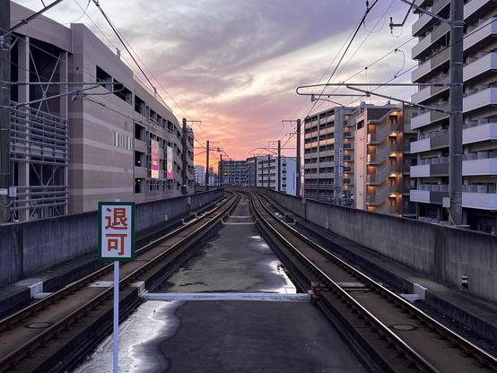 A sunset view along railway tracks, flanked by modern buildings on either side. A sign in the foreground features the characters "退可". Everything is bathed in a pinkish light.