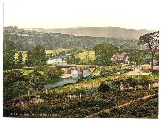 The image portrays a picturesque rural landscape featuring a charming bridge. The setting is likely in England, as indicated by the text on the print "Torrington Rothern Bridge." This old photograph captures an idyllic countryside scene with rolling hills adorned with lush greenery and scattered trees providing depth to the background.

In the foreground, there's a vineyard or cultivated field with neatly trimmed bushes lining its boundaries. A dirt path runs through this area, suggesting it may be used for walking or small vehicles. The central focus is on an old stone bridge crossing over a tranquil river that reflects the sky and surrounding landscape. This structure appears to have multiple arches.

On either side of the stream are houses with visible architectural details such as gabled roofs and chimneys, indicative of residential buildings from this era. A fence runs along one edge of the property adjacent to the vineyard or field, possibly demarcating private land.

The overall atmosphere is serene and untouched by modernity, providing a glimpse into rural life in late 19th-century England. The colors are vivid with varying shades of green indicating different types of vegetation, while brown tones dominate the structure of both the bridge and houses.