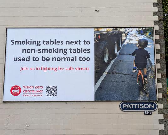 Ein Foto einer Plakatwand, auf deren rechter Hälfte ein Kleinkind auf einem Fahrrad neben den Reifen eines LKW dargestellt ist. Links steht auf Englisch "Smoking tables next to non-smoking tables used to be normal too".