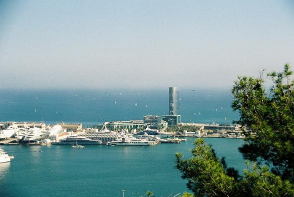 A wide-angle photograph of Barcelona’s port and coastline, taken from Montjuïc. The image captures a panoramic view of the harbor, filled with docked yachts and boats. The water is a deep blue-green, contrasting with the lighter blue of the sky. In the background, the city’s skyline is visible, including a tall, modern skyscraper with a cylindrical shape. The foreground features green trees, framing the left side of the image. The lighting suggests daytime, with clear visibility and no visible clouds.
